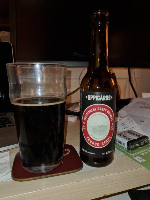 En stout-öl i glas bredvid en ölflaska från Oppigårds bryggeri på ett skrivbord.