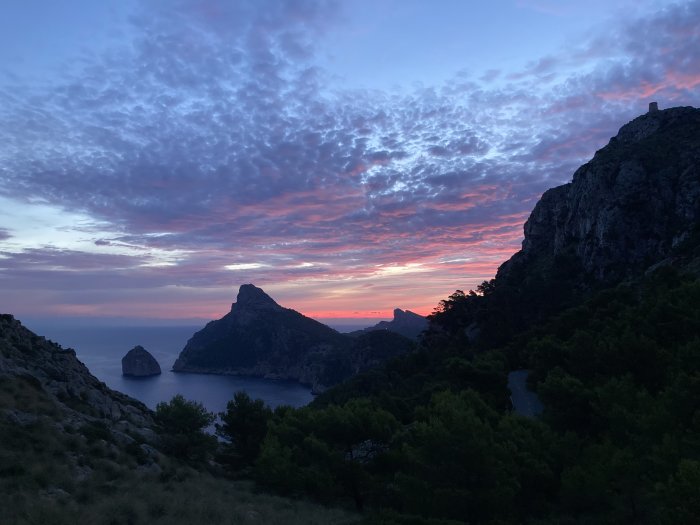 Soluppgång med dramatiska rosa och blå himmel över Cap de Formentor med silhuetter av berg och träd.