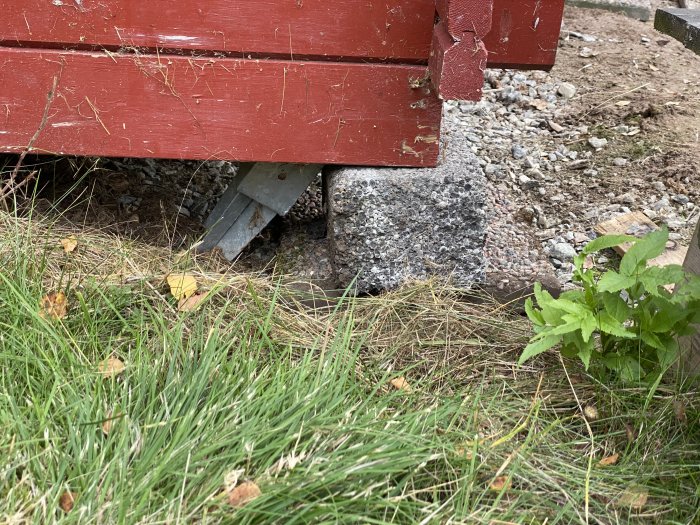 En del av en röd stuga med temporär upplyftning på plankbitar och lecablock, belägen på en grusad slänt.