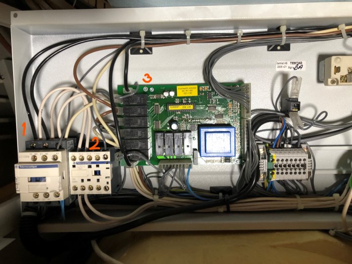 Elektriskt skåp med komponenter: kontaktorer märkta 1 och 2, kretskort med reläer och etikett för testning.