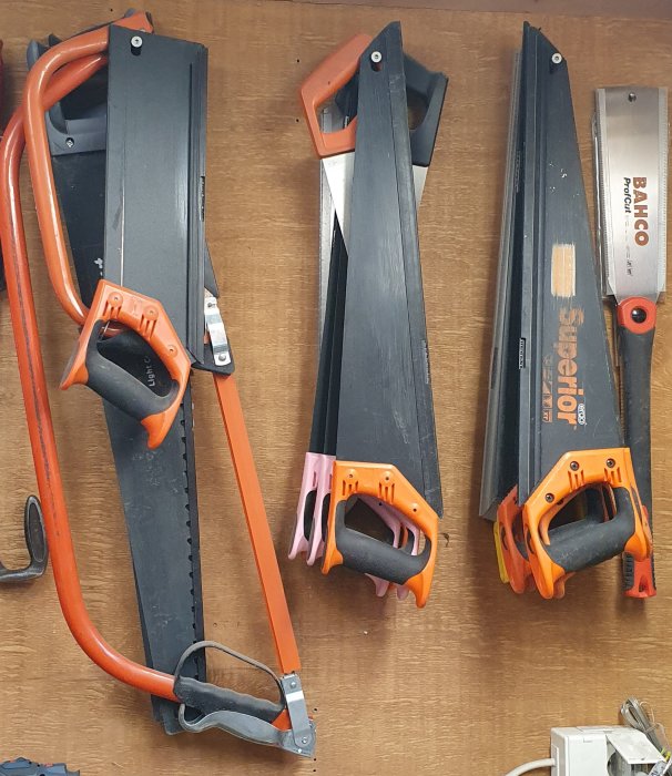 Fyra handsågar med orange handtag upphängda på en verktygstavla.