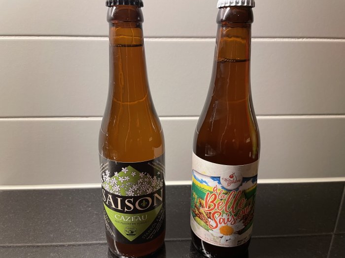 Två ölflaskor av saison-typ står på en köksbänk framför vita kakelplattor.