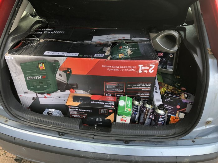 Baklucka på bil full med byggvaror och verktyg, inklusive hyvel i trasig kartong, batteriladdare och motorolja.