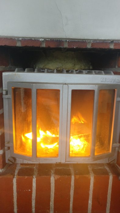 Eldstad med brinnande spiskassett och synlig isolering runt skorströret ovanför.