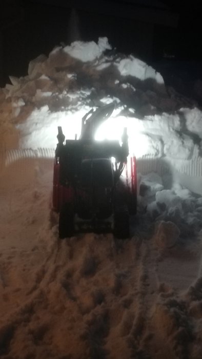 Snöslunga i arbete på natten med upplyst snöhög framför.