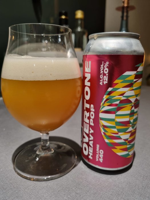 Ölglas med skumkrona bredvid en färgglad ölburk på en bordsyta.