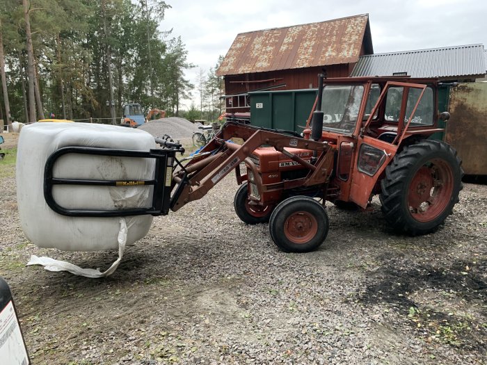 Äldre röd traktor med frammonterad balgrip utomhus på en grusgård med lantlig miljö i bakgrunden.
