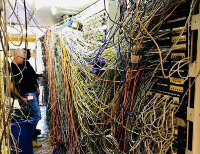 Rörigt serverrum med trassliga kablar och en förbryllad person i bakgrunden.