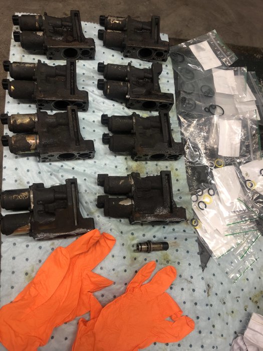 Flera demonterade och smutsiga byggnadsdelar på en verkstadsgolv med orange handskar och nya delar i påsar.