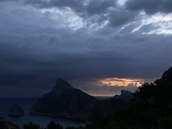 Dimmig solnedgång bakom berg vid kust med molnigt himmel.