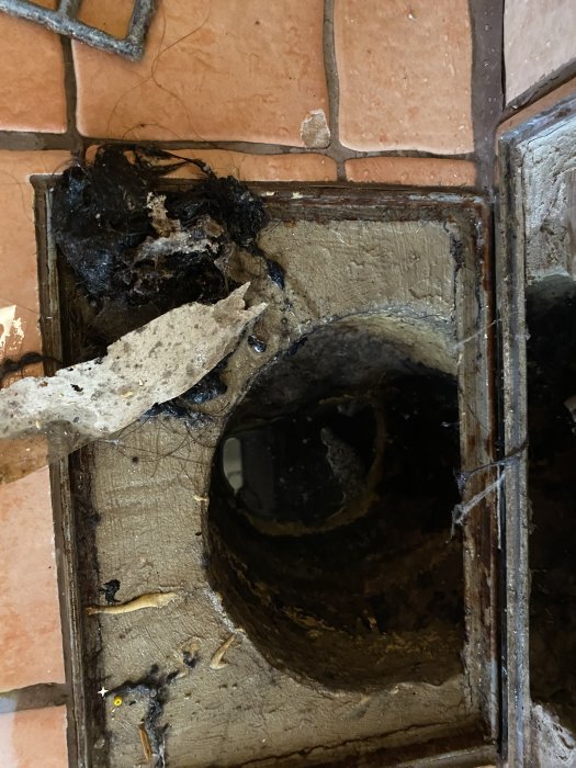 Öppen golvbrunn med synliga rörsystem och smuts, omgiven av terrakottaklinker som behöver renoveras.