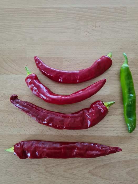 Fem röda chili-frukter och en grön på träyta, stark färgpigmentering, redo att bli flakes.