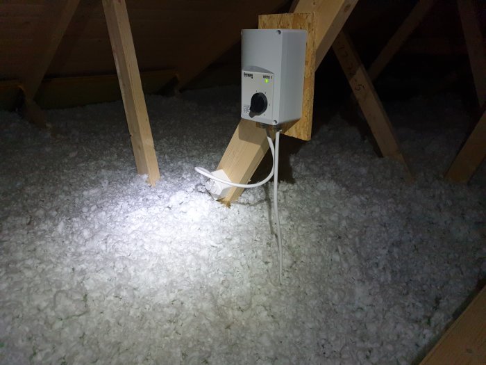 Kanalfläkt på vinden omgiven av lösullsisolering, delvis belyses av en ficklampa.