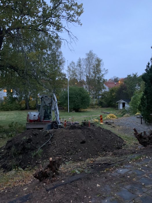 Grävmaskin och uppgrävd jord i en trädgård för byte av vattenmätare 3.5 meter under mark.