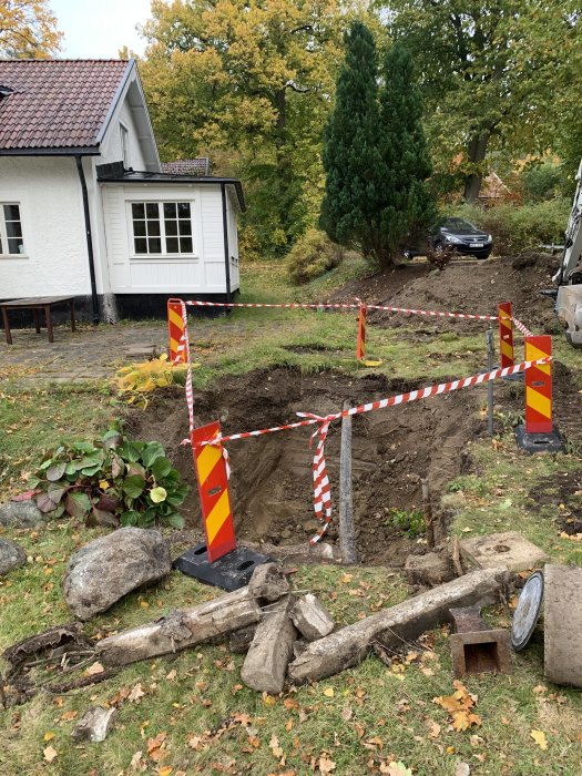 Avgrävning i en trädgård med säkerhetsstaket och grävmaskin i bakgrunden vid ett hus.