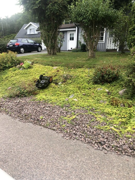 Gräsklippare som välte på en gräsbevuxen slänt framför ett hus med parkerad bil.