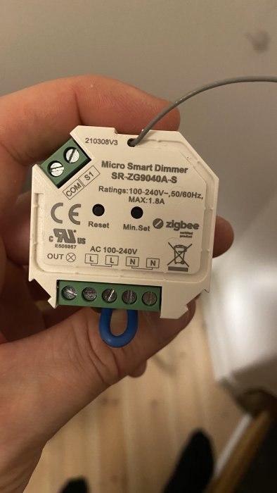 Hand som håller en ZigBee Micro Smart Dimmer med synliga terminaler och kabelanslutningar.