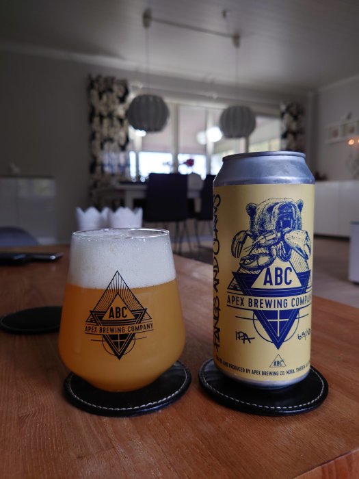 Ölglas fyllt med öl bredvid en ölburk på ett bord med suddig inredning i bakgrunden.