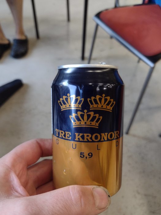 Hand håller en burk öl märkt "Tre Kronor" med alkoholstyrka 5,9% i en inomhusmiljö.
