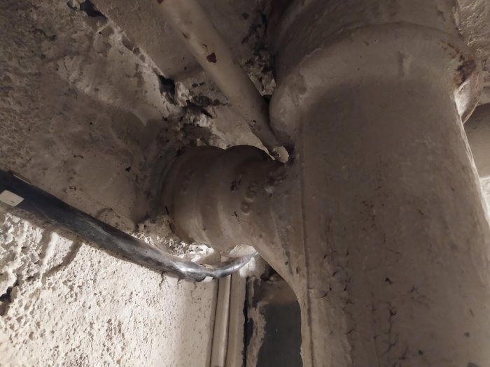 Sprucken muff på ett vertikalt rör med ett anslutet horisontellt rör i en betongmiljö, troligt luftningsrör i badrum.