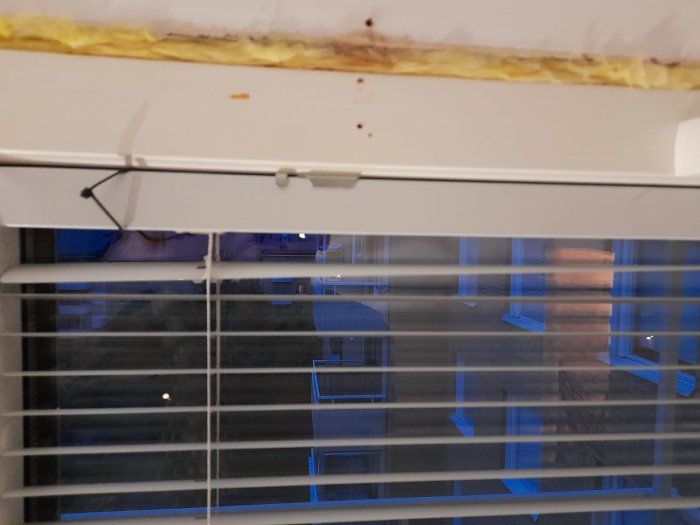 Öppen vit persienn med oviss fästanordning på ett fönster, synlig rost vid fönsterkarmen.