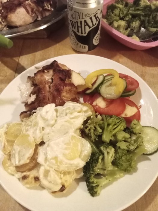 Tallrik med baconlindad kycklingfilé, potatisgratäng, grönsaker och ölburk i bakgrunden.