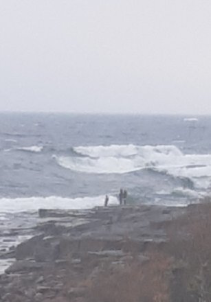 Två personer står på klippor vid havet med vågor i bakgrunden.
