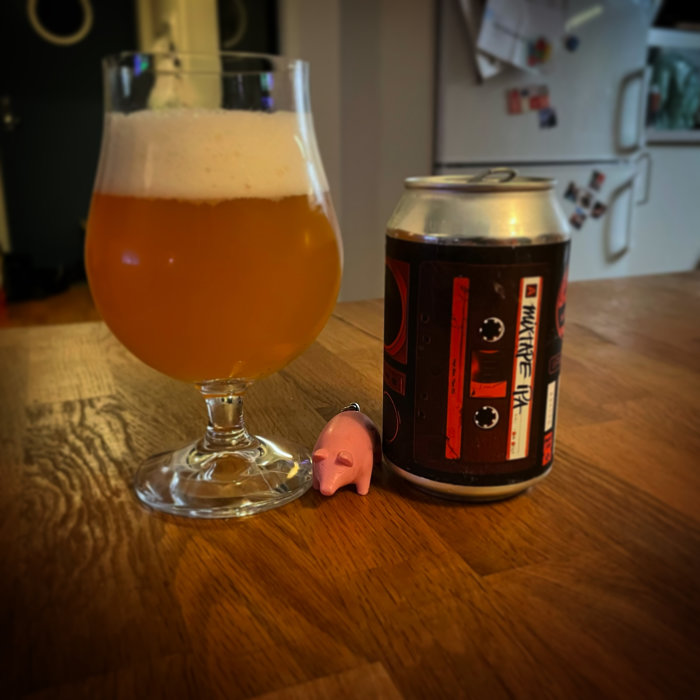 Ölglas med skummande öl bredvid en burk märkt "Pwrkörk" och en liten rosa plastgris på ett träbord.