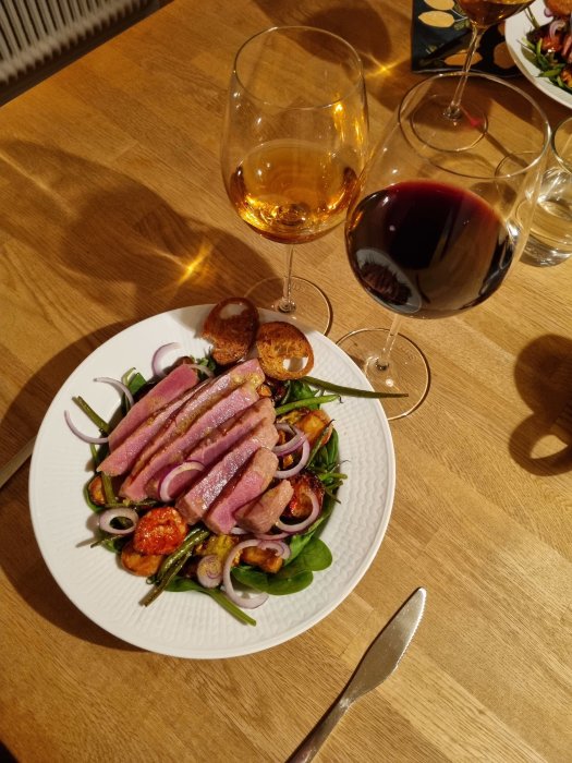 Grönsallad med skivor av rosa tonfisk, gröna bönor, rödlök, och halverade tomater, serverat med vitt och rött vin.