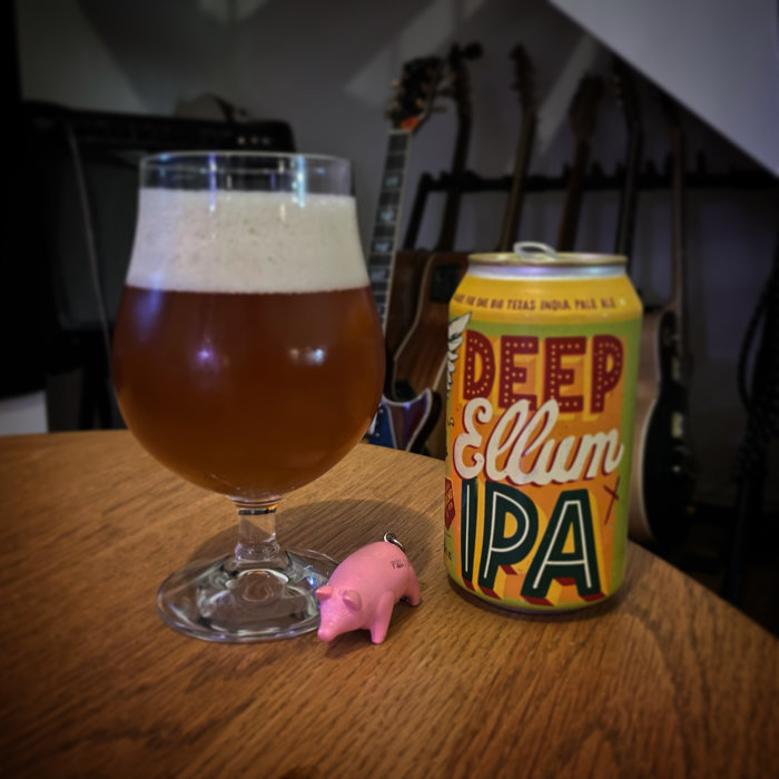 Ett glas öl bredvid en burk Deep Ellum IPA och en liten rosa plastgris på ett bord, med gitarrer i bakgrunden.