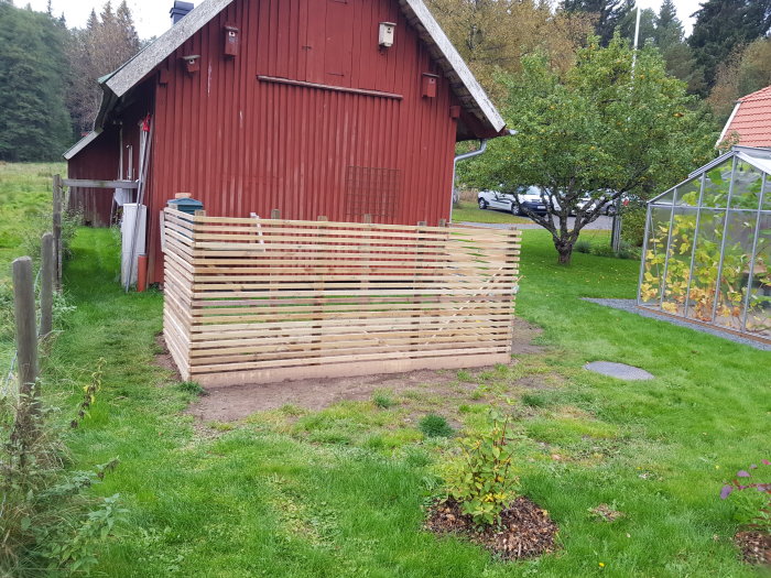 Nybyggd trädgårdskompost i trä framför ett rött lantligt hus och ett växthus.