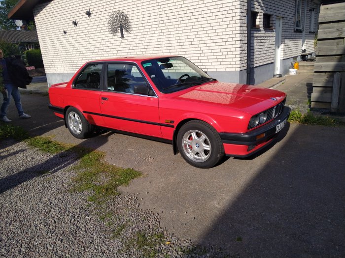 Röd BMW 325 E30 parkerad framför en byggnad, dagtid.