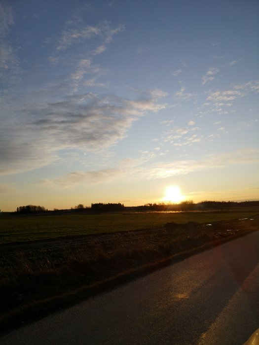 Solnedgång över ett öppet fält med delvis molnig himmel och väg i förgrunden.