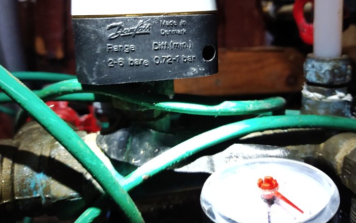 Närbild på en tryckströmbrytare märkt "Danfoss" i ett blött pumprum, omgiven av rör och kablar.
