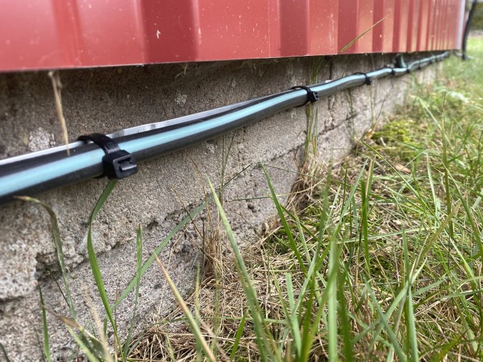 Elektriska kablar fästa med svarta buntband på aluminiumskena längs husgrund.