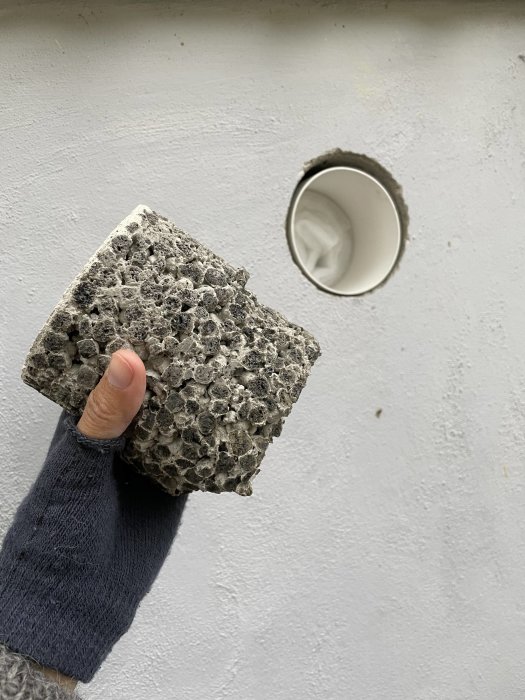 Hand i stickad tröja håller betongbit bredvid ett plastventilationsrör i en vägg.