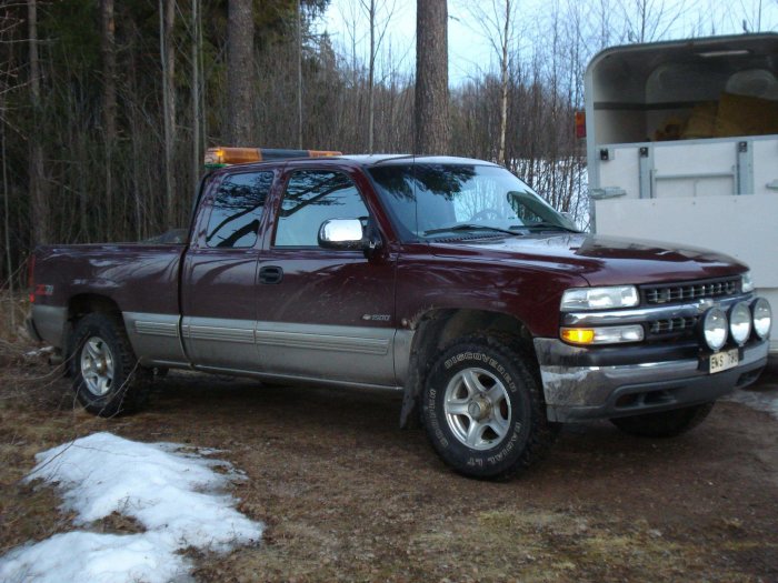 Stor pickup truck parkerad utomhus, delvis på gräs med snöfläckar och träd i bakgrunden.