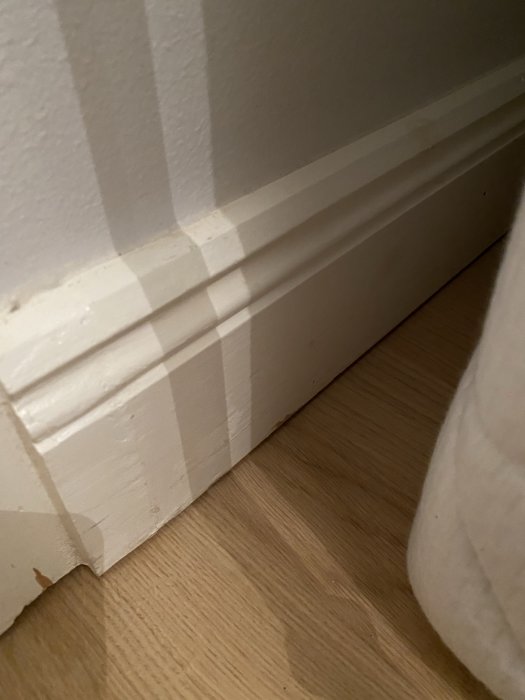 Valkoinen jalkalista seinän alareunassa ja puulattia niiden alla; huoneen sisänurkka.