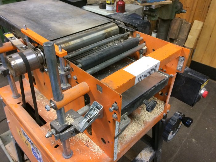 Orange planhyvel med synliga rullar och reglage, saknar fast bord, spån syns.