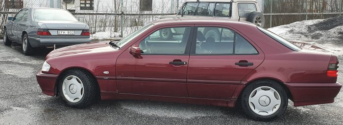 Röd bil parkerad på grus med en silverfärgad bil i bakgrunden, snöfläckar runtomkring.