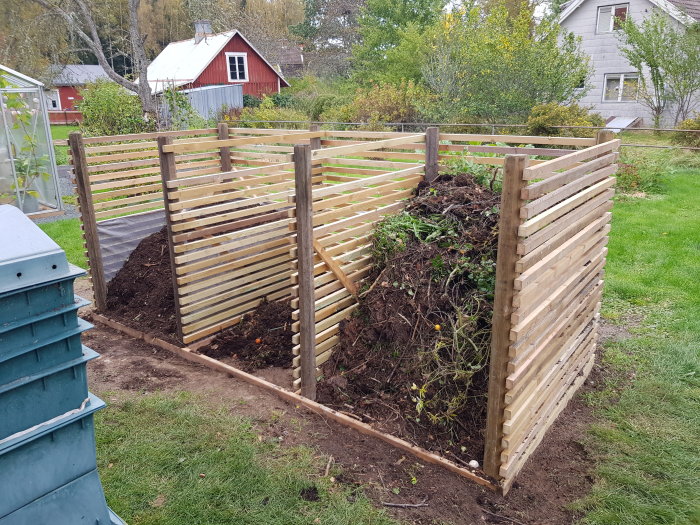 Nybyggd träkompost i trädgården full med kompostmaterial, med röd stuga i bakgrunden.