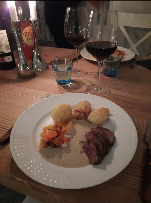 Middag med rådjurssadel, hasselbackspotatis, pepparsås och rött vin på bord med ljus.