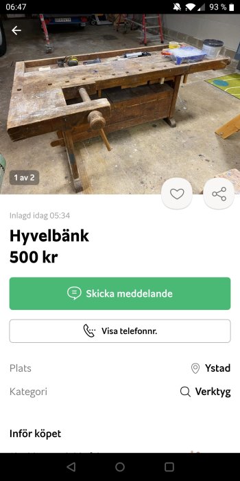 Använd hyvelbänk i trä med skruvstäd, verktyg och byggmaterial i bakgrunden i en verkstad.