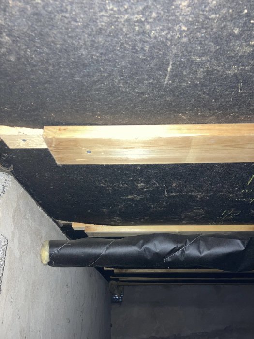 Undervy av ett otillräckligt isolerat golvbjälklag med synliga träreglar och svart plastfolie.