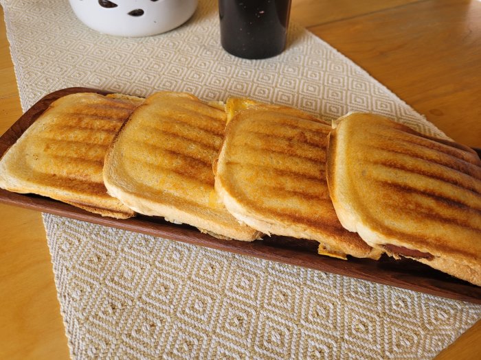 Fyra grillade mackor på en skärbräda med mönstrad kökshandduk i bakgrunden.