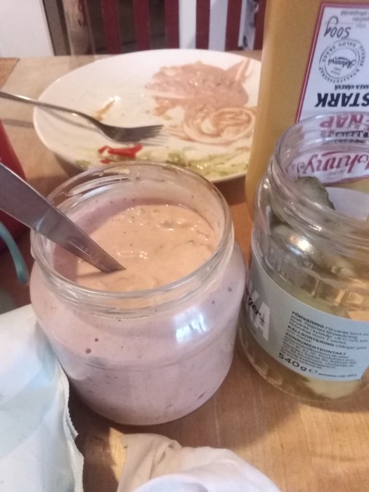 Hemmagjord rosa dressing i glasburk, inlagda gurkor i burk och rester av maträtt på tallrik.