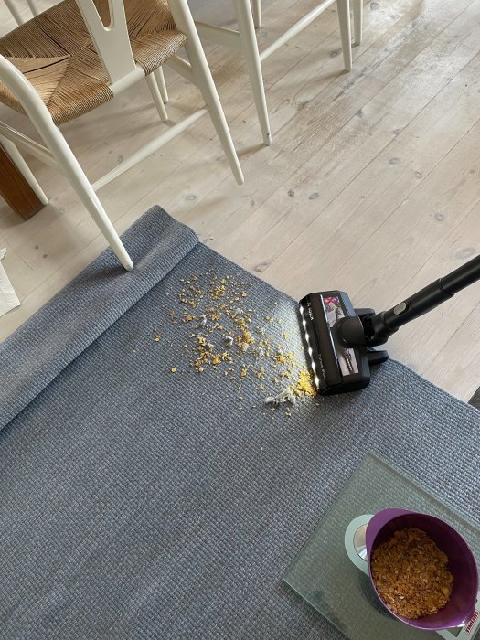 Dammsugare bredvid en hög med uppstädade smulor på en grå matta, med en stol i bakgrunden.