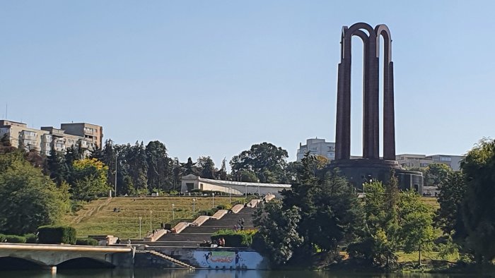 Stort rödbrunt monument vid flod med omgivande grönska och gångväg under blå himmel.