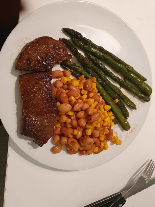 Grillad ryggbiff med sauterade sparrisstjälkar och bönsalsa med majs på en vit tallrik.
