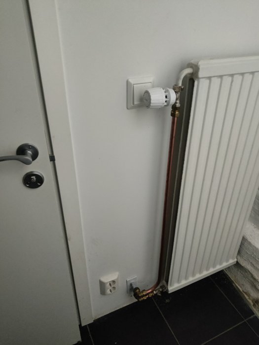 Termostat ovanför en elkontakt och rörarbeten som korsar varandra på ett oorganiserat sätt vid ett element.